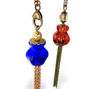 May include: Two necklaces featuring decorative pendants. One has a cobalt blue glass bead, a gold-tone hammered cap, and a copper chain tassel. The other has a translucent red glass bead, a clear bead with colorful flecks, and a dark metal chain tassel.