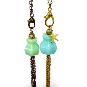 May include: Two necklaces featuring glass bottle pendants. The left pendant is pale green with a floral bead and copper chain tassel. The right pendant is light blue with a gold-tone starfish charm, a ribbed bead, and a gold-tone chain tassel.