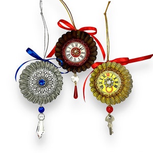May include: Three Christmas ornaments shaped like clock faces with different colored backgrounds. The ornaments are silver, red, and gold. Each ornament has a different design and a different type of embellishment. The silver ornament has a blue jewel in the center, the red ornament has a red jewel, and the gold ornament has a clear jewel.
