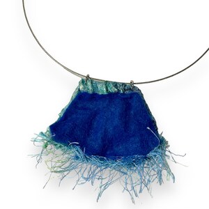 May include: A blue and green fabric necklace with a fringe detail. The necklace is made of a soft, textured fabric and has a simple, elegant design.
