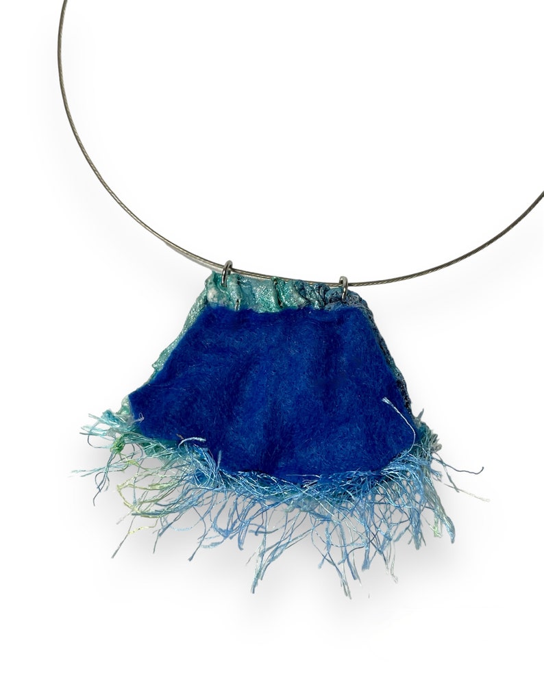 May include: A blue and green fabric necklace with a fringe detail. The necklace is made of a soft, textured fabric and has a simple, elegant design.
