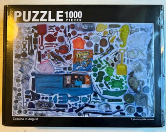 Può includere: Un puzzle da 1000 pezzi con un collage di vari oggetti in vari colori. Il puzzle include un sacchetto di Cheetos, un cavalluccio marino giallo e una tenda blu. Il puzzle è intitolato "Coquina in August".