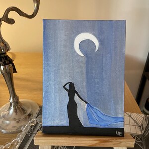 May include: A painting on canvas of a woman in a long dress with a blue scarf blowing in the wind. The background is a blue sky with a white crescent moon.