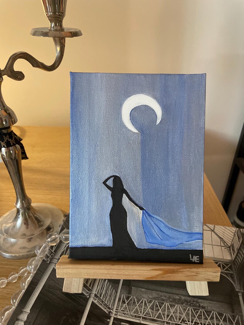 May include: A painting on canvas of a woman in a long dress with a blue scarf blowing in the wind. The background is a blue sky with a white crescent moon.