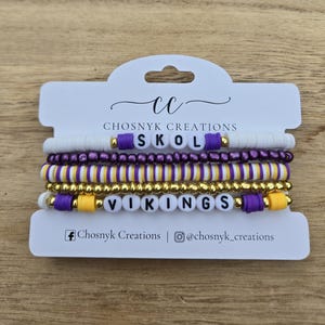 Pode incluir: Pilhas de pulseiras de contas em branco, roxo, dourado e amarelo. As pulseiras soletram "SKOL" e "VIKINGS" com contas de letras brancas. O cartão diz "Chosnyk Creations" com identificadores de mídia social.