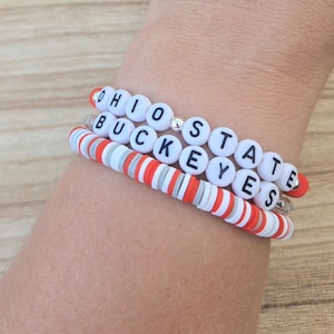May include: Three bracelets stacked on a wrist. The top bracelet has white letter beads spelling "OHIO STATE" with a silver bead. The middle bracelet spells "BUCKEYES". The bottom bracelet is red, white, and gray striped.