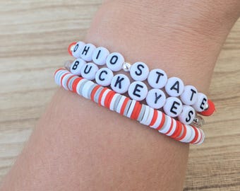 May include: Three bracelets stacked on a wrist. The top bracelet has white letter beads spelling "OHIO STATE" with a silver bead. The middle bracelet spells "BUCKEYES". The bottom bracelet is red, white, and gray striped.