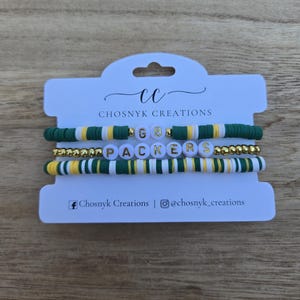 May include: Four handmade bracelets with green, yellow, white, and gold beads. One bracelet spells out "PACKERS" with white letter beads. The other bracelet has the letters "GB" with gold beads. The bracelets are displayed on a white card with the text "Chosnyk Creations".