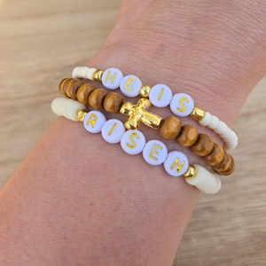 May include: A set of three bracelets with white beads and gold lettering spelling "He is Risen" with a gold cross charm.