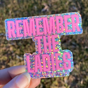 May include: A holographic sticker with the text "REMEMBER THE LADIES" in bright pink block letters. The sticker has a glittery, iridescent border and is held against a blurred outdoor background.