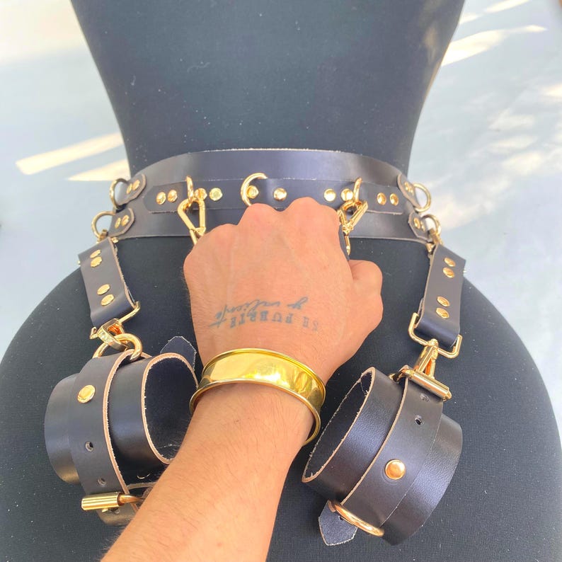 May include: Black leather restraint set with gold-colored hardware, including a belt, wrist cuffs, and connecting straps. The set is displayed on a black mannequin. A gold bracelet is also visible.