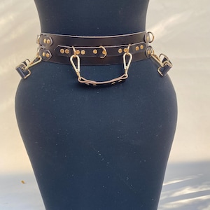 May include: A dark brown leather belt with gold-colored hardware. The belt features a series of small gold rivets, D-rings, and clasps. A small handle is attached to the front of the belt. The belt is displayed on a black mannequin.