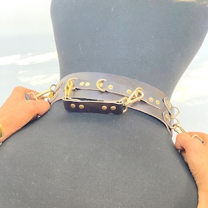 May include: A dark brown leather collar with gold-colored hardware, including clasps and rivets. The collar is displayed on a black mannequin, showcasing its design and construction. The image highlights the craftsmanship and details of the accessory.