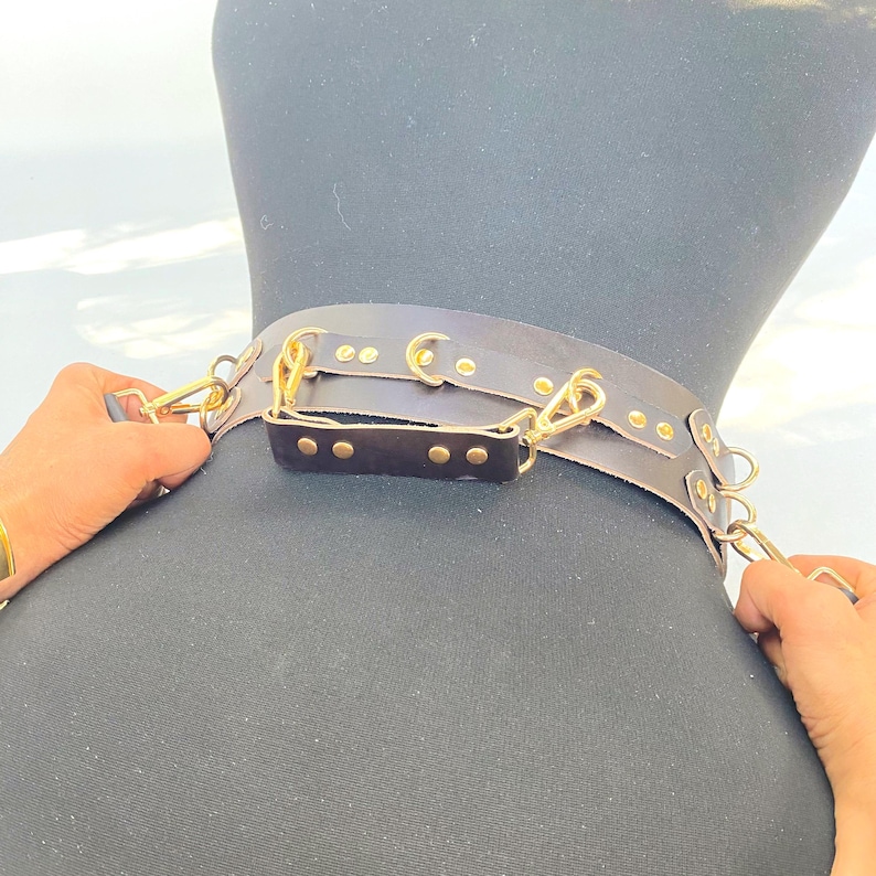 May include: A dark brown leather collar with gold-colored hardware, including clasps and rivets. The collar is displayed on a black mannequin, showcasing its design and construction. The image highlights the craftsmanship and details of the accessory.