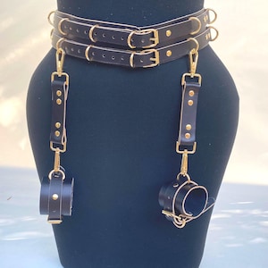May include: Black leather body harness with gold-colored hardware. The harness features two belts, suspender straps, and wrist cuffs. The cuffs and straps are connected with gold-colored clasps and rivets. The harness is displayed on a black mannequin.