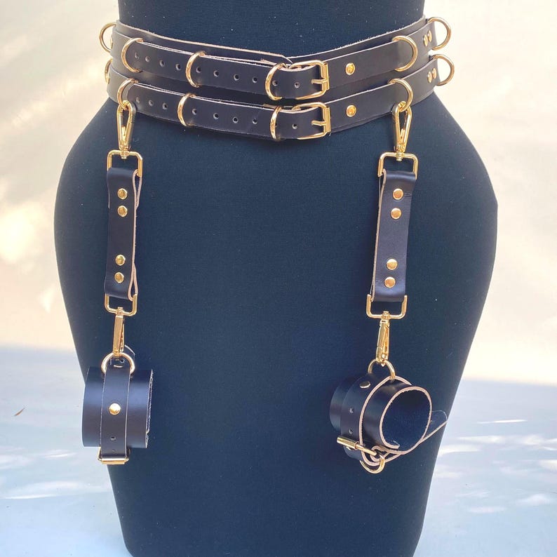 May include: Black leather body harness with gold-colored hardware. The harness features two belts, suspender straps, and wrist cuffs. The cuffs and straps are connected with gold-colored clasps and rivets. The harness is displayed on a black mannequin.