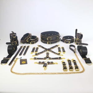 May include: A collection of black leather restraints with gold-colored hardware, including cuffs, collars, and harnesses. The set includes a leash and a gold chain. The items are arranged on a white surface.