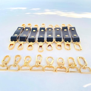 May include: A collection of black leather key fobs with gold-colored hardware, including snap closures and swivel clasps. The key fobs are arranged in two rows against a white background, showcasing their design and craftsmanship.