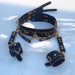 May include: Black leather restraint set with gold-colored hardware. The set includes a collar, wrist cuffs, and connecting straps. The collar and cuffs are secured with buckles and rivets. The hardware includes D-rings and clasps.