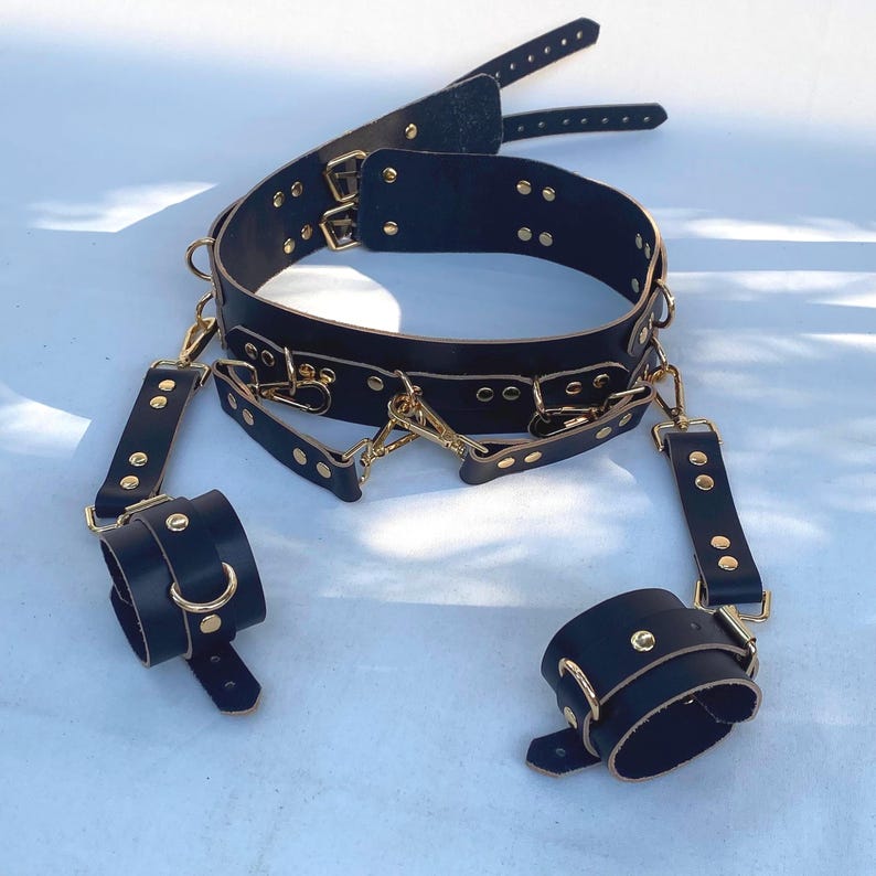 May include: Black leather restraint set with gold-colored hardware. The set includes a collar, wrist cuffs, and connecting straps. The collar and cuffs are secured with buckles and rivets. The hardware includes D-rings and clasps.