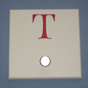May include: White square sign with a red capital letter "T" and a white round light switch.
