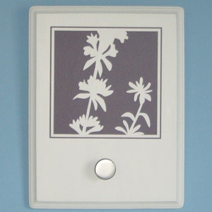 May include: White ceramic wall mounted switch plate with a floral design in a light purple frame. The switch plate has a round, white knob.