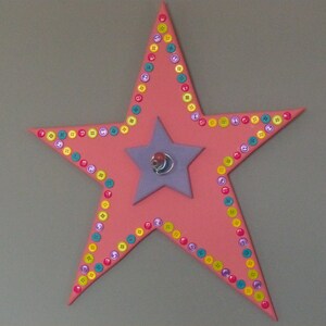 May include: A pink star-shaped wall decoration with a smaller purple star in the center. The star is decorated with colorful buttons.