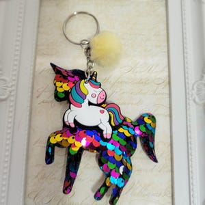 May include: A vibrant unicorn keyring with a small, fluffy yellow pom-pom. The keyring features a large unicorn with a sequined body in various colours, and a smaller unicorn riding on its back. The background is a framed print.