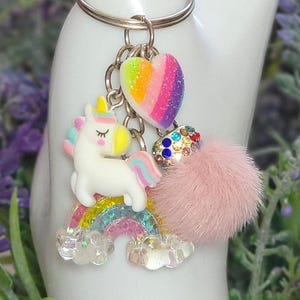 May include: A silver keychain with a collection of charms. The charms include a white unicorn with a rainbow mane and tail, a rainbow heart, a pink fluffy ball, and a rainbow-colored arch with clouds.