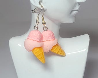 May include: A pair of dangle earrings featuring miniature ice cream cones. Each earring has a light pink ice cream scoop atop a yellow waffle cone. The earrings are suspended from silver-colored fish hook ear wires.