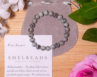 May include: A Kiwi Jasper bracelet with gray and black speckled beads, resting on a gray pouch. The bracelet is displayed with a card that reads "Kiwi Jasper" and "SHELBEADS Jewellery Boutique."