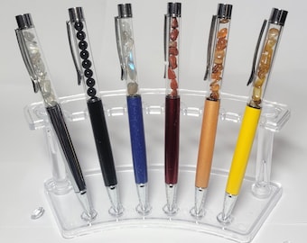 May include: Six pens with clear barrels filled with stones and beads. The pens have silver accents and are displayed in a clear plastic holder. The pens are black, blue, red, orange, and yellow.