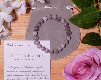 Puede incluir: Un brazalete de cuentas de turmalina rosa sobre una bolsa gris, con una tarjeta y flores. El brazalete presenta cuentas de color púrpura claro. La tarjeta dice "Pink Tourmaline" y "SHELBEADS Jewellery Boutique". Una rosa rosa y flores blancas también están en la imagen.
