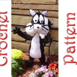 May include: Crocheted stuffed animal toy of Pepe Le Pew, a cartoon skunk, in black and white. The toy is standing on a bed of brown mulch with green plants and pink flowers in the background.