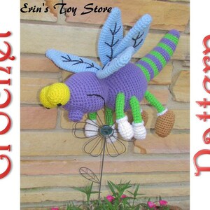 May include: A crocheted purple dragonfly with green and white stripes, yellow eyes, and blue wings. The dragonfly is perched on a metal flower with a brown stem.