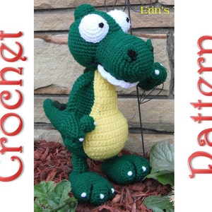 Puede incluir: Un juguete de cocodrilo de crochet verde y amarillo con ojos y dientes blancos. El juguete está de pie sobre una cama de mantillo marrón.