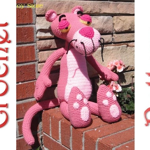 Puede incluir: Juguete de peluche rosa tejido a crochet del personaje de dibujos animados La Pantera Rosa. El juguete está sentado en una pared de ladrillo con plantas verdes en el fondo.