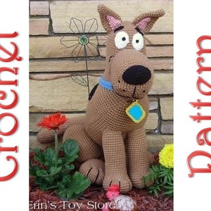 Puede incluir: Peluche de Scooby Doo tejido a crochet sentado frente a una pared de ladrillo. El peluche es marrón con un collar azul y una etiqueta azul. El juguete está sentado en un lecho de mantillo marrón con plantas verdes y flores.