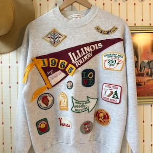 May include: A light gray sweatshirt adorned with various patches, including a pennant that reads "Illinois Tollway 1965." Other patches feature text like "Cub Day Camp," "Lake View," and "Kansas." The sweatshirt has a crew neck.