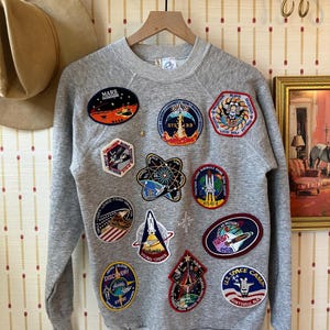 May include: A gray crewneck sweatshirt adorned with numerous colorful embroidered patches. The patches feature space-themed designs, including rockets, planets, and the NASA logo. The sweatshirt is hanging on a wooden hanger.