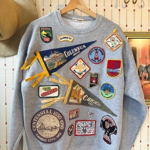 May include: A gray sweatshirt adorned with numerous embroidered patches. Patches feature text like "Columbus Ohio", "Chicago Ill", and "Kansas City Mo". The patches showcase various designs, including pennants, landmarks, and logos, creating a vintage aesthetic.