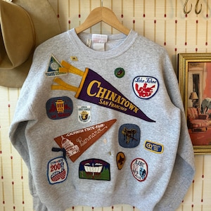 May include: A gray sweatshirt adorned with various patches, including a purple pennant that reads "Chinatown San Francisco." Other patches feature text like "Lake Tahoe Love You" and "California State Fair." The sweatshirt has a crew neck.