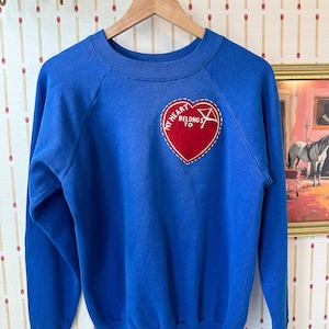 以下が含まれることがあります： 長袖とクルーネックのロイヤルブルーのスウェットシャツ。胸には「MY HEART BELONGS TO」の文字と白いグラフィックが入った赤いハート型のパッチが縫い付けられています。スウェットシャツは木製のハンガーに掛けられています。