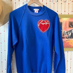 Op de afbeelding: Een koningsblauwe sweatshirt met een ronde hals en lange mouwen. Een rood hartvormig embleem met de tekst "MY HEART BELONGS TO" is op de linkerborst genaaid. De sweatshirt hangt aan een houten hanger.