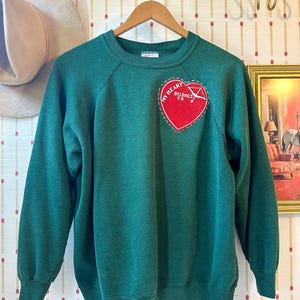 May include: A forest green long-sleeve sweatshirt featuring a red heart-shaped patch on the upper left chest. The patch includes white stitched text that reads MY HEART BELONGS TO and a white graphic of an arrow piercing the heart shape.