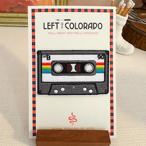 May include: A cassette tape patch with a rainbow stripe design, displayed on a card with the text "LEFT OF COLORADO" and "HELL-BENT AND WELL-DRESSED." The card is propped up by a small wooden stand. The text "REPAIR, PERSONALIZE, KEEP" is also visible.