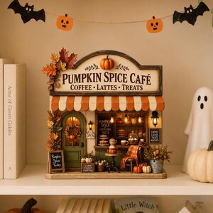 May include: A miniature Pumpkin Spice Café shop display featuring a sign that reads Pumpkin Spice Café Coffee Lattes Treats, an orange and white striped awning, a green door, autumn foliage, miniature pumpkins, and interior lighting.