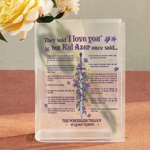 May include: A clear acrylic bookend featuring a quote from the book "The Powerless Trilogy" by Lauren Roberts. The quote reads "They said "I love you" but Kai Azer once said..." and is followed by a list of 12 statements. The bookend features a purple and green floral design with a sword.