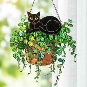 Op de afbeelding: Een gebrandschilderde hangende plantenbak met een zwarte kat die op weelderig groen blad rust. De plantenbak heeft de vorm van een bruine mand en is opgehangen aan een ketting. De kat heeft gele ogen en de bladeren hebben verschillende groentinten.