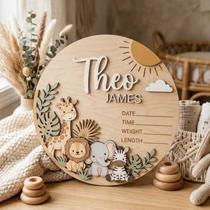 May include: A circular wooden birth announcement sign featuring the name Theo James, a sun, a cloud, and cut-out illustrations of a giraffe, lion, elephant, and zebra. The sign includes printed lines for date, time, weight, and length details.